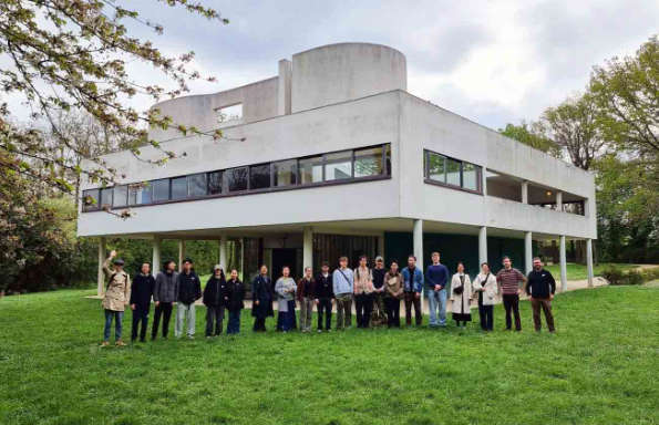 Modernist building surrounded by trees with people standing on a grass lawn in front.