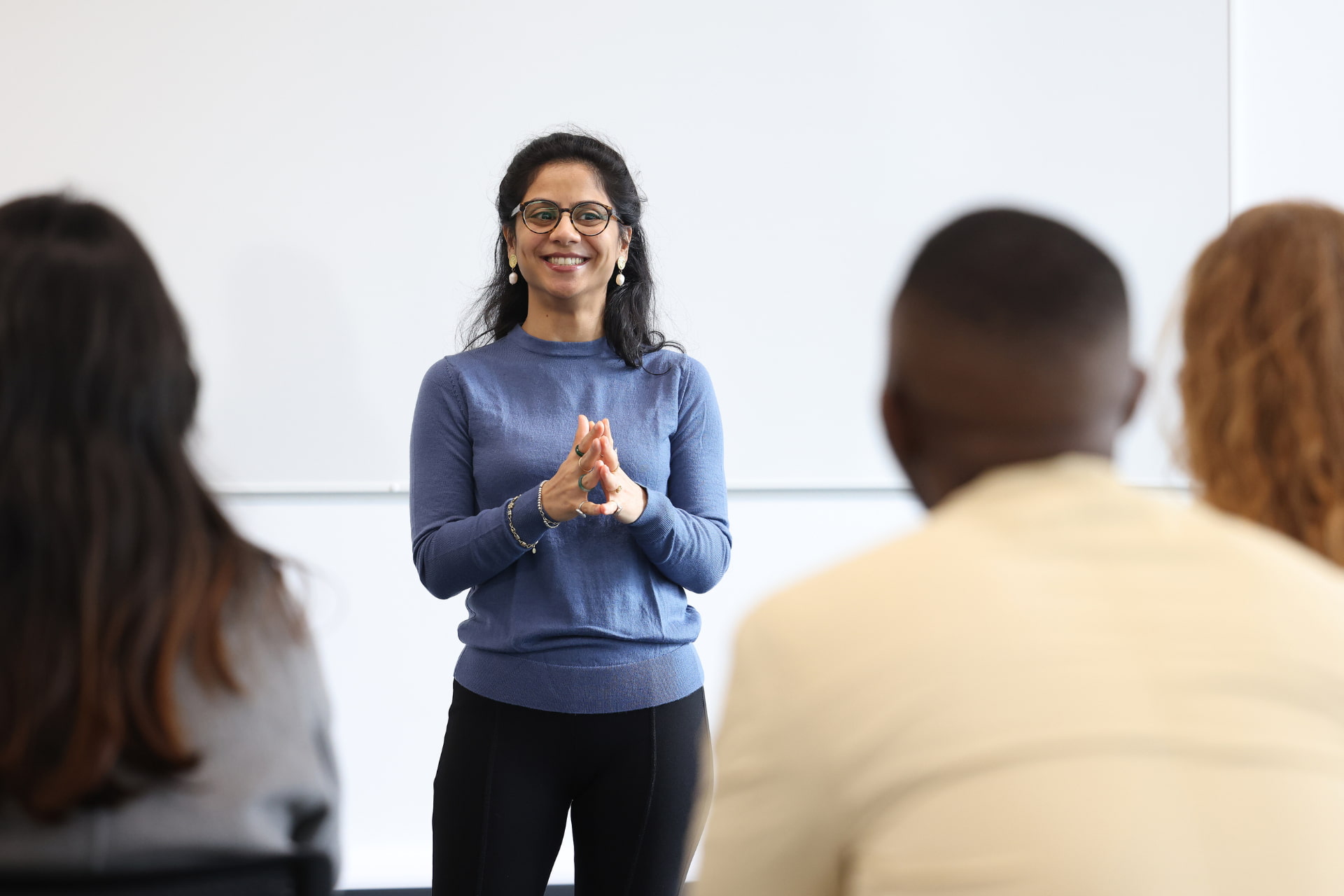 A smiling woman standing at the front of a classroom. In the foreground you see the backs of three students.