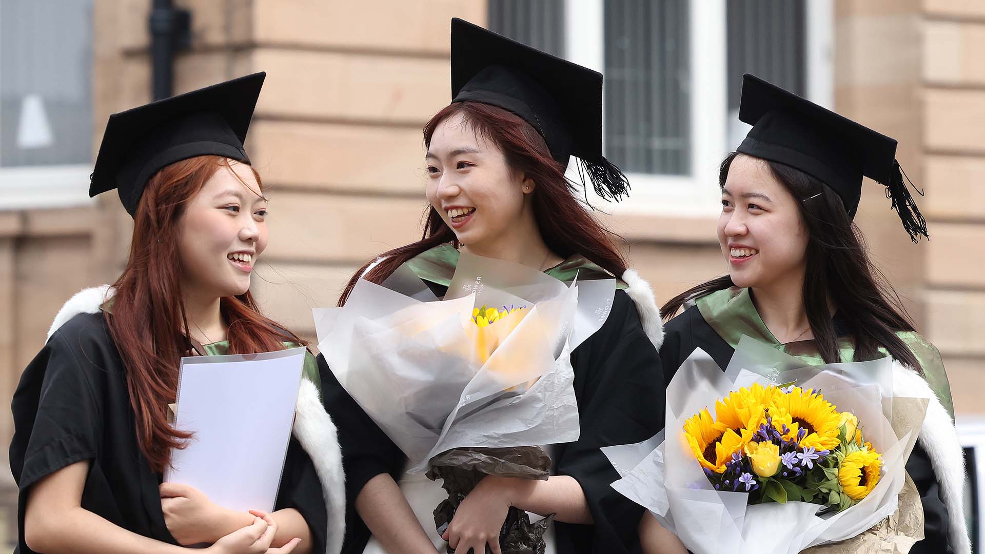 Three graduates wearing academic dress and holding flowers