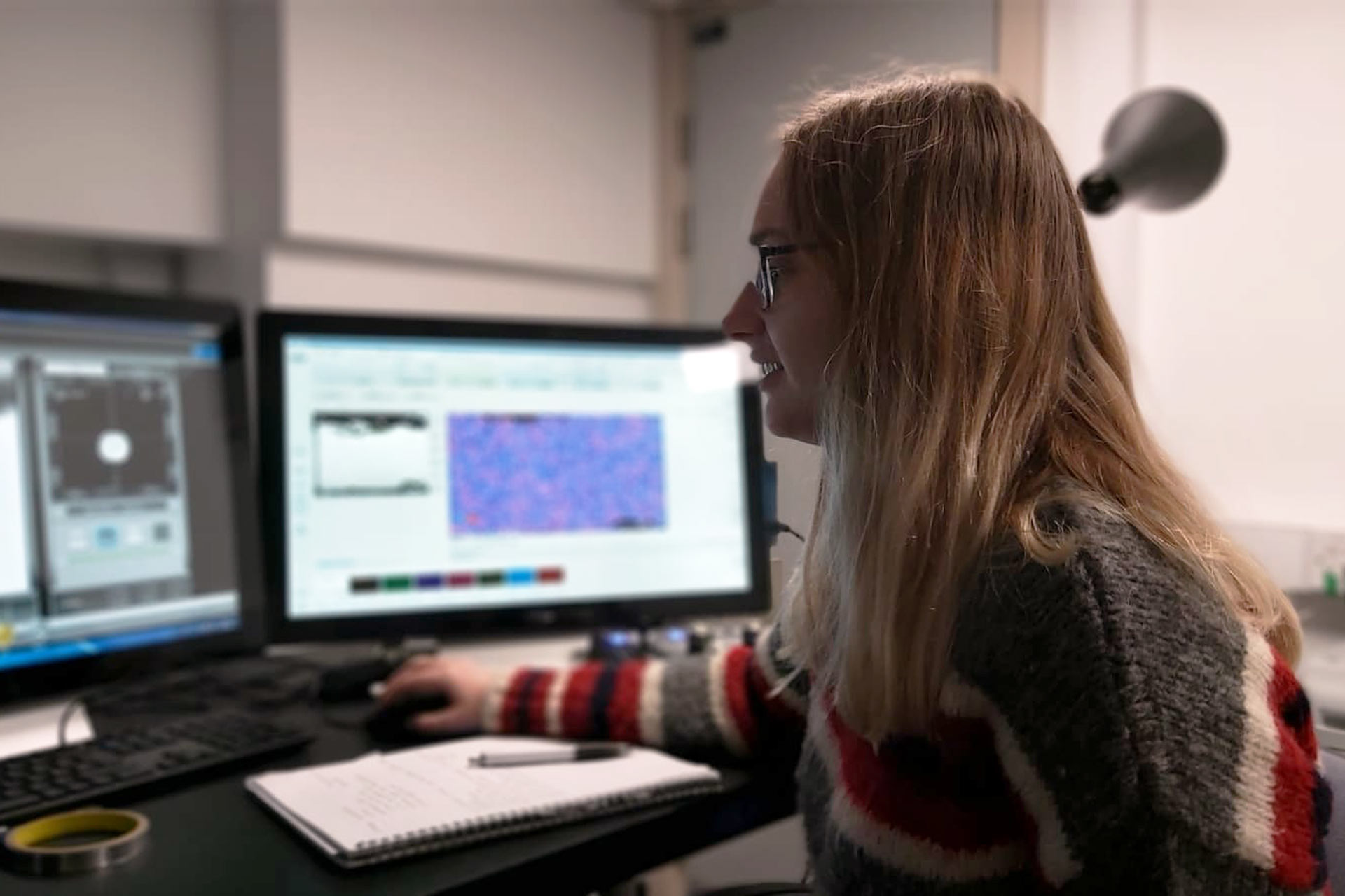 A woman with long blonde hair sat in front of two computer screens using the mouse