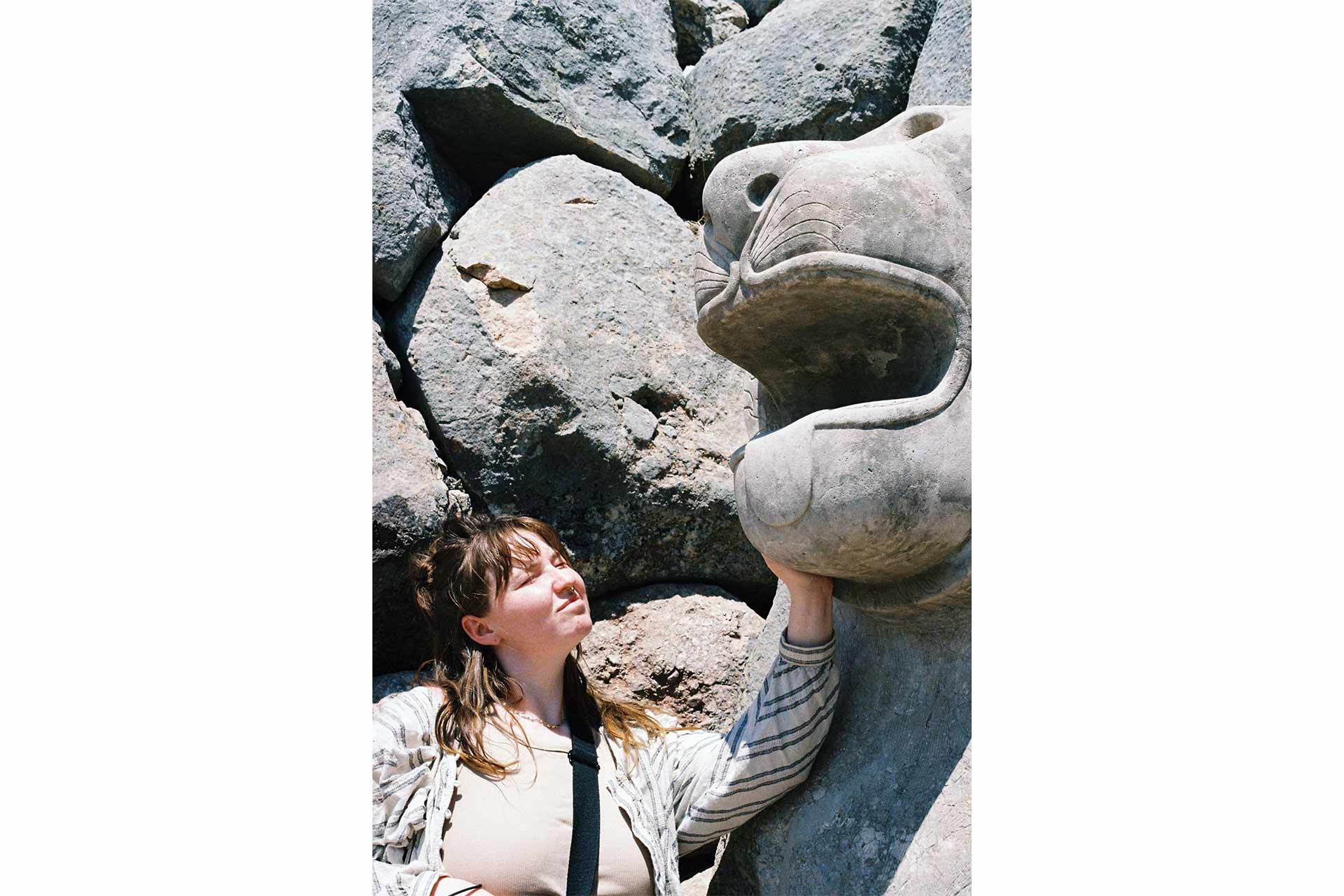 A stone statue of a lion on the right, with a young woman to the left of it holding her hand up to the mouth of the statue