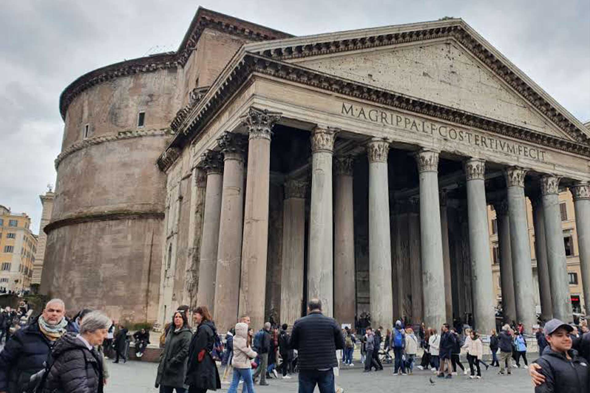Large ancient building with large columns and Roman writing on the front, a large number of people gathered outside of it