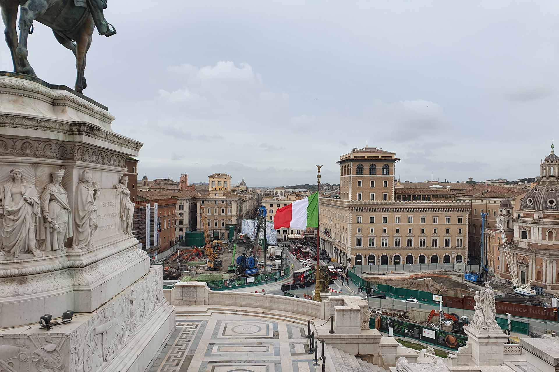 Looking down on the view of a city, an Italian flag in the middle and old Italian buildings in the background, on the left is a statue of a horse, the rest cropped out of the picture