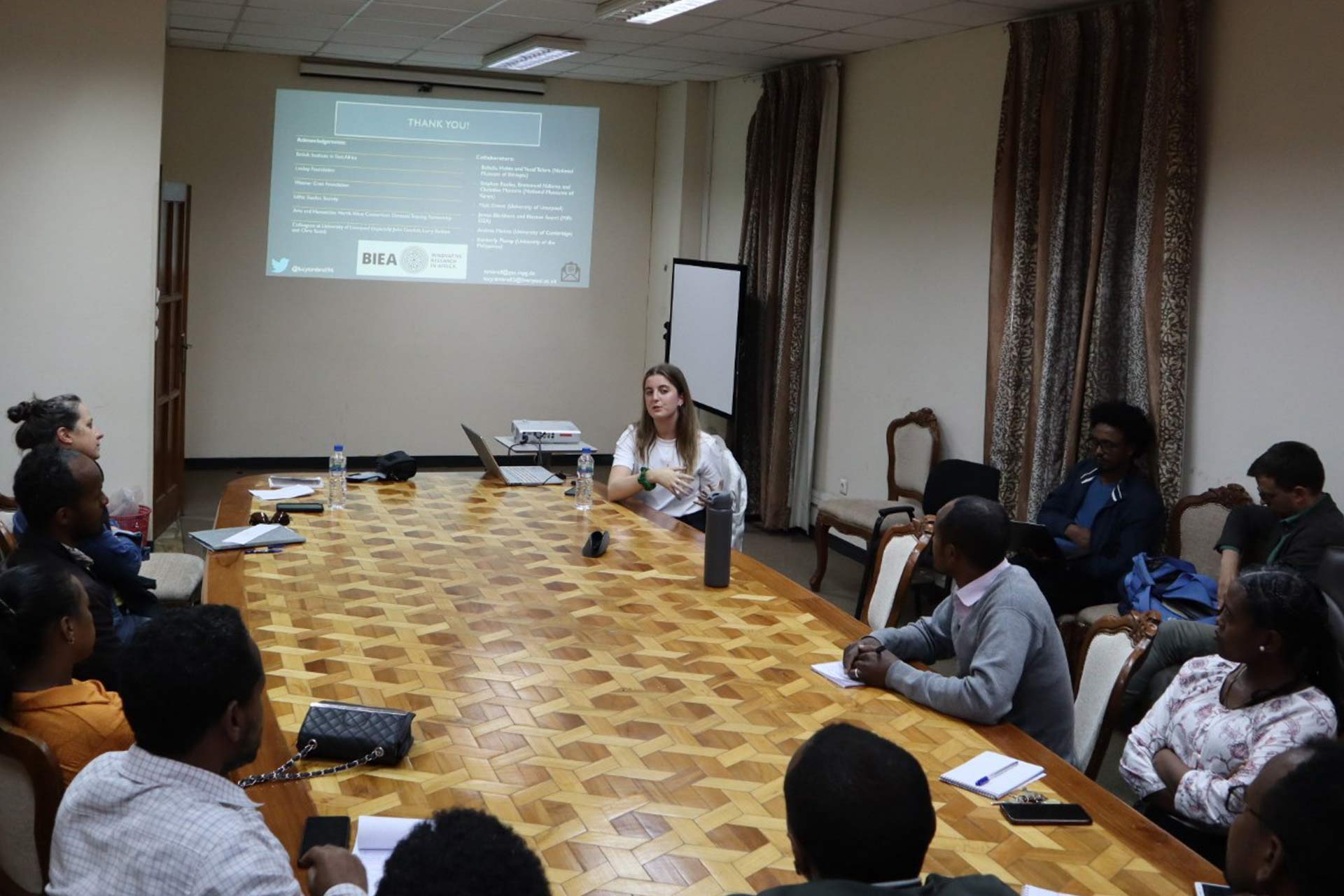 10 people sitting around a large table whilst a woman presents in front of a projector screen