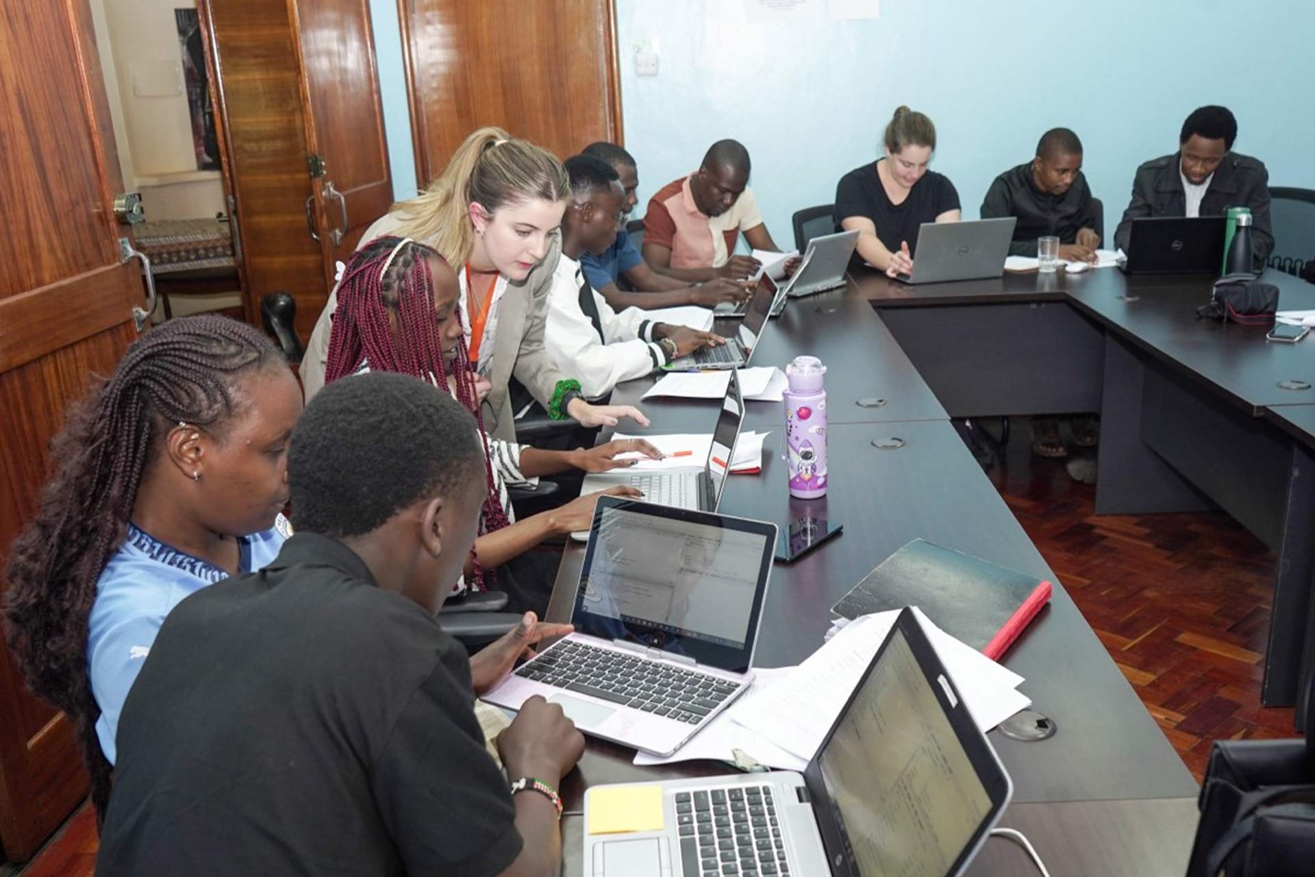 9 people sitting down intently working on their laptops whilst a woman stands over and looks at their work