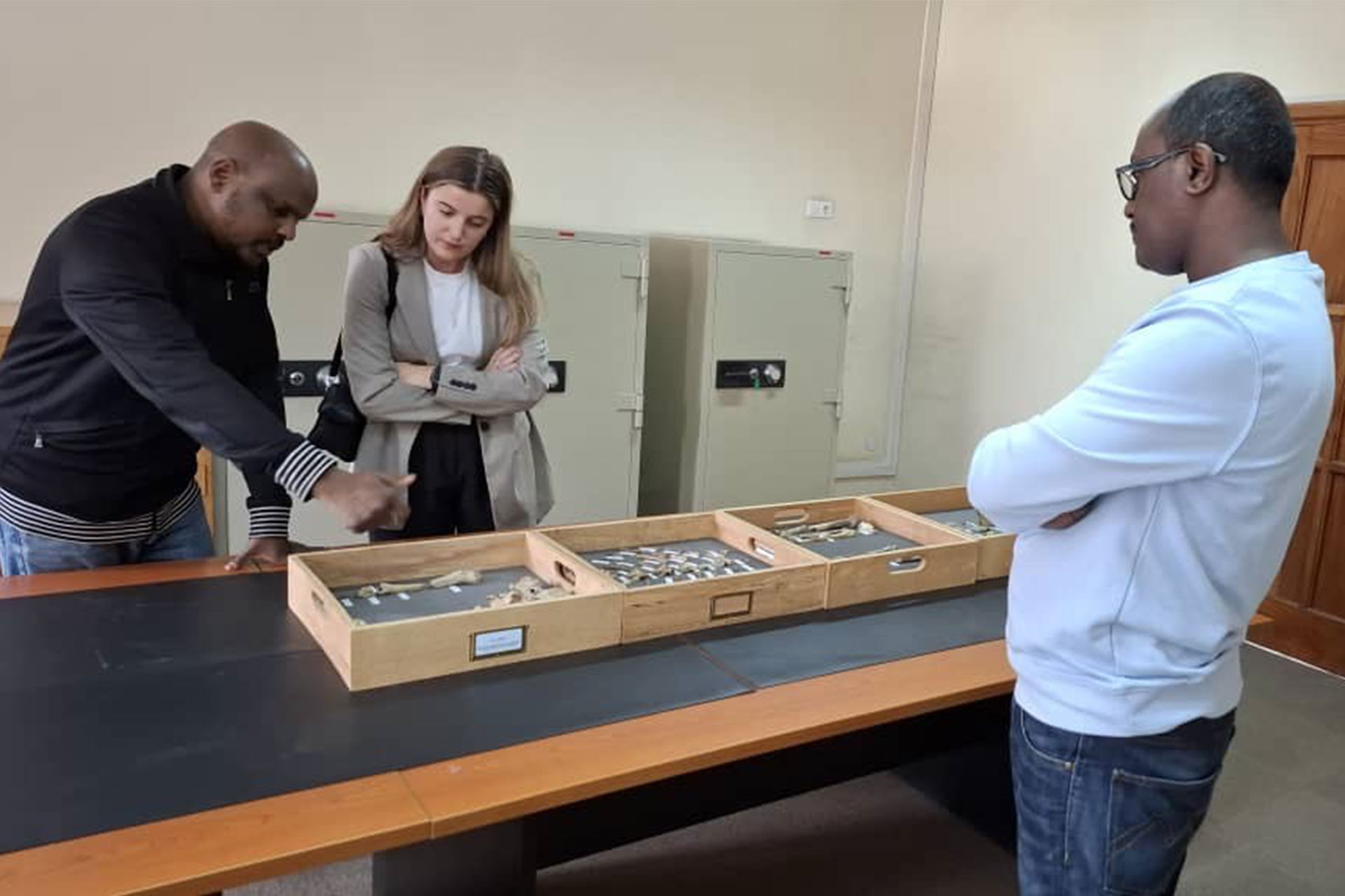 Three people standing over a display cabinet with remains/bones, one man points towards a specific bone while the others look interestedly at the remains.