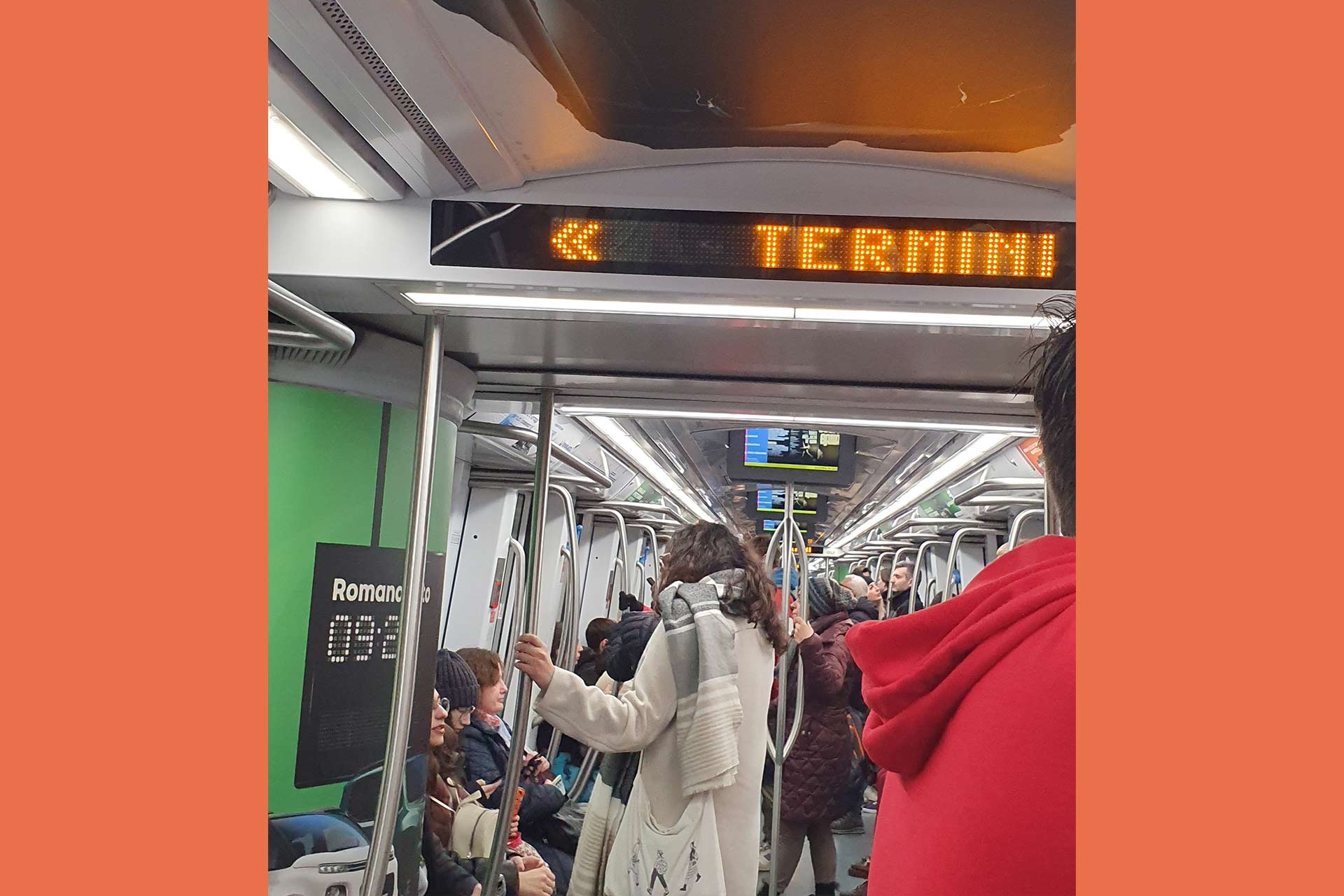 The inside of a train or subway carriage, people sitting and standing, the digital display reads 'Termini'