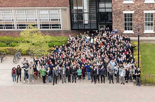 Large group of people standing out side a building looking up at a camera.