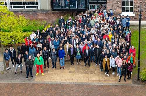 Large group of people standing out side a building looking up at a camera.