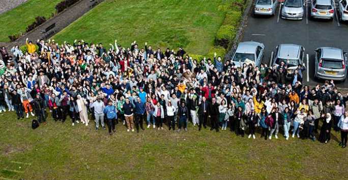 Group of several hundred people standing on grass next to a car park. The photograph is taken from several hundred feet up.