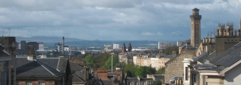 View across the slated rooftops of a Scottish city, a shipbuilding crane is visible in the background, hills are visible on the horizon.