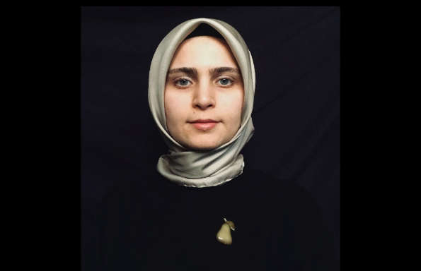 Young woman dressed in black wearing a silver hijab, with a bronze apple broach on her lapel.