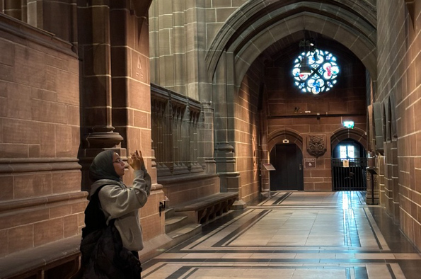 Young woman standing in a corridor in a cathedral holding a camera. The wallls are made from sandstone and there is a door with a stained glass window above it at the end.