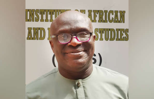 Smiling bald middle aged black man wearing glasses and a collarless shirt. They are standing outside in front of a sign reading Institute for African and Diaspora Studies.