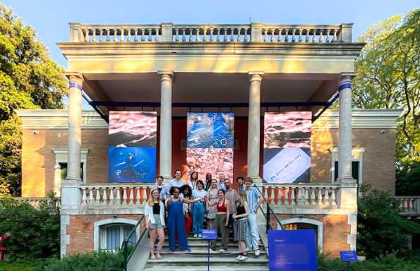 A group of people standing on stone steps leading up to a classical brick and stone pavilion. Behind are mature trees.