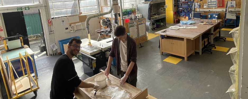 Two men in a large spacious workshop standing at a bench building an architectural model. One is smiling at the camera.