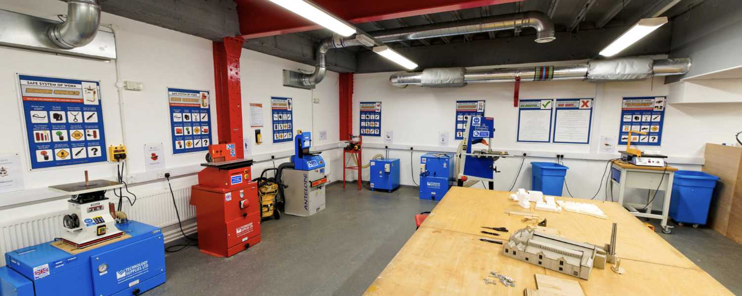 Workshop space with a large wooden table in the middle of the room. Around the walls are a number of woodworking machines, health and safety posters and extractor equipmentor.