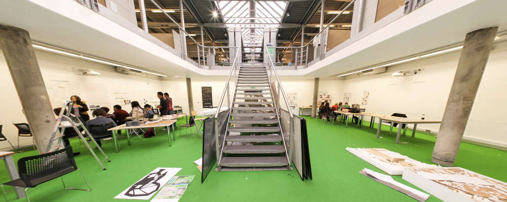 Inside a modern hi-tech studio with a steel frame and pitched glass roof. In the middle is a steel staircase, students can be seen working and presenting design schemes.