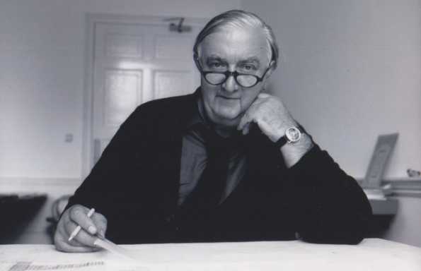 Older, slightly balding man with glasses wearing a shirt, tie and cardigan, sitting at a desk holding a pen whist looking at a drawing.