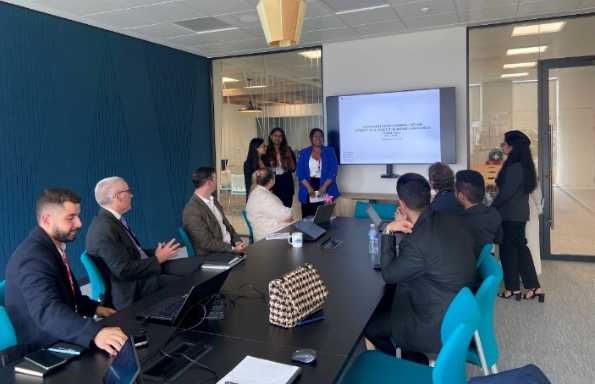 A meeting room, 8 men and women sit around a table looking at a screen showing a presentation. On the right hand side beside the screen three women stand taking to the room.