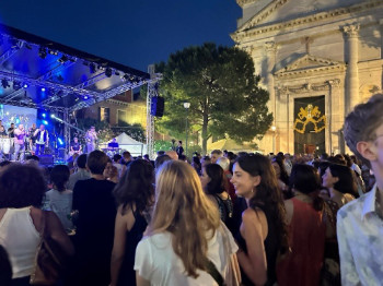 A group of people enjoying an outdoor concert. The stage has trees and a classical building to one side.