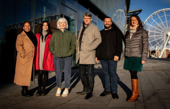 Four women and two men standing in front of the glass facade of of conference centre in the winter sunshine. Behind them is a large ferris wheel.