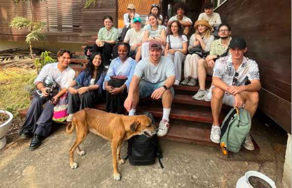 Group of students sitting on a set of wooden steps outside a wooden building. In front of them is a thin stray dog.