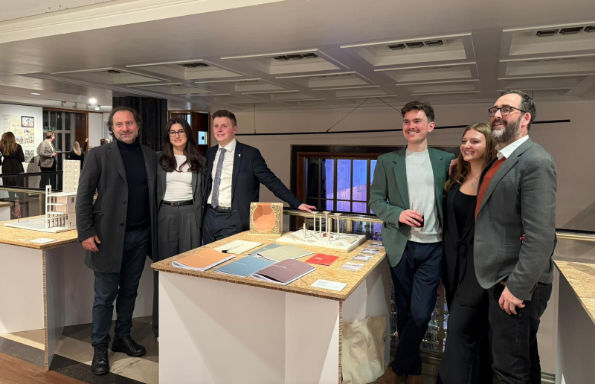 Two women and four men stand proudly around a table. On the table are a series of brochures and two architectural models.