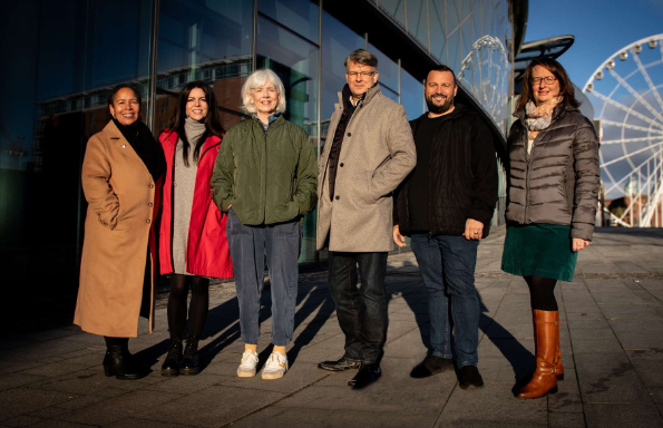 Four women and two men standing in front of the glass facade of of conference centre in the winter sunshine. Behind them is a large ferris wheel.