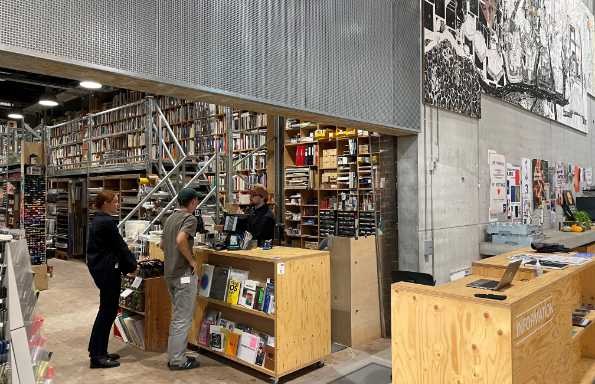 A woman and a man are being served by a man stabding at a wooden counter. There are artist materials and books for sale on extensive shelving. Behind the counter is a short set of steps leading up to more bookshelves.