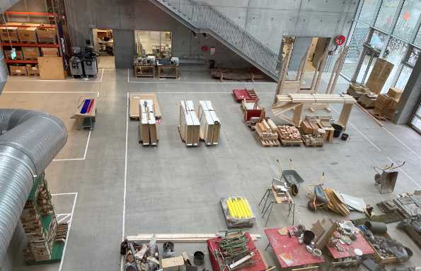 Concrete floor of a large open plan workshop. There are wooden crates and a wheelbarrow in the middle, tables with tools, glass doors at one end and a set of concrete stairs run up the far wall.