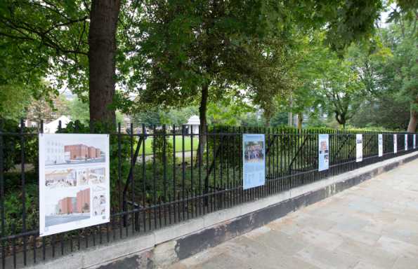 Posters attached to park railings, behind is a hedge and trees overhanging, in front a stone pavement.