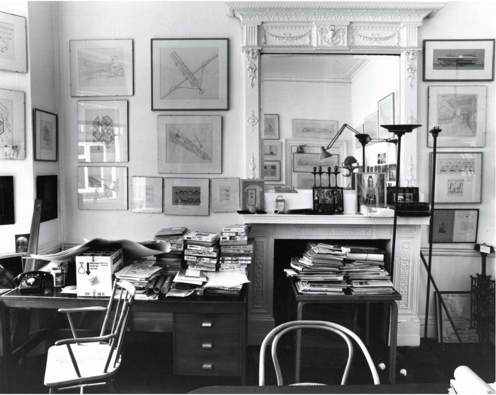 A cluttered rooom with an ornate fireplace and mirror. A desk and table in front are pilled high with books and papers. Architectural drawings cover the walls.