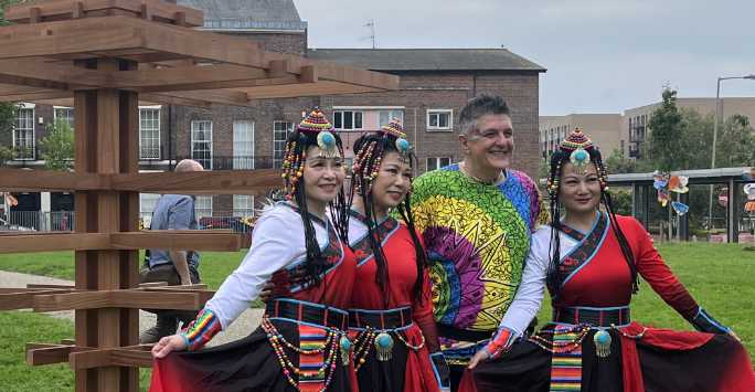 Four people, three in traditional Chinese dress stand outside in front of a tall wooden block sculpture.