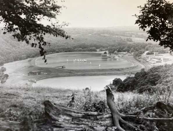 Vintage photograph of a small reservoir, with boats sailing. It is surrounded by woodland. In the foreground is a pile of cut wood.