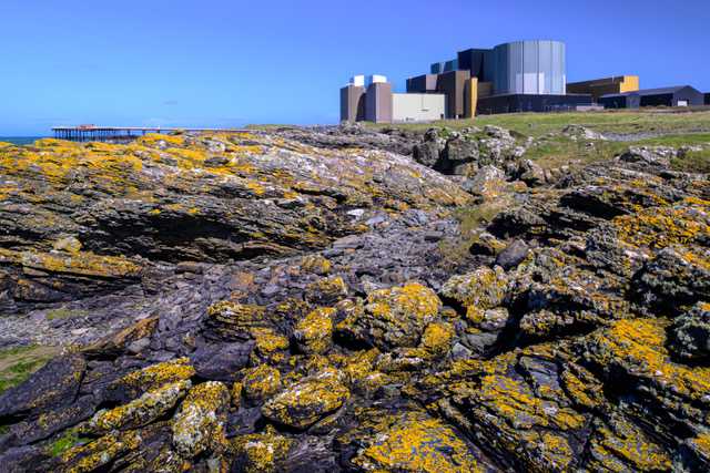 An outcrop of rock covered in lichen, in the background grass land with a Nuclear Power station behind.