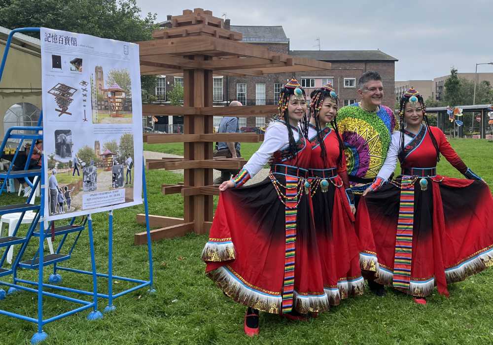 Four people, three in traditional Chinese dress stand outside in front of a tall wooden block sculpture.