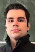 head and shoulder view of a young white man with short dark slicked back hair. wearing a jacket with a turned up collar.