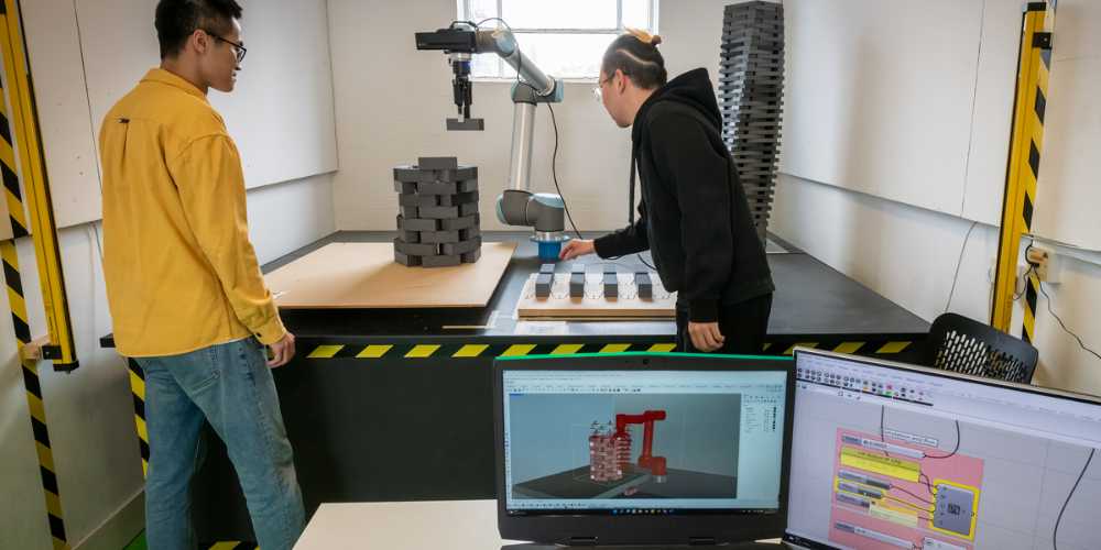 Swo students standing in front of a robot arm. The robot arm is building a structure. In the foreground are two laptops.