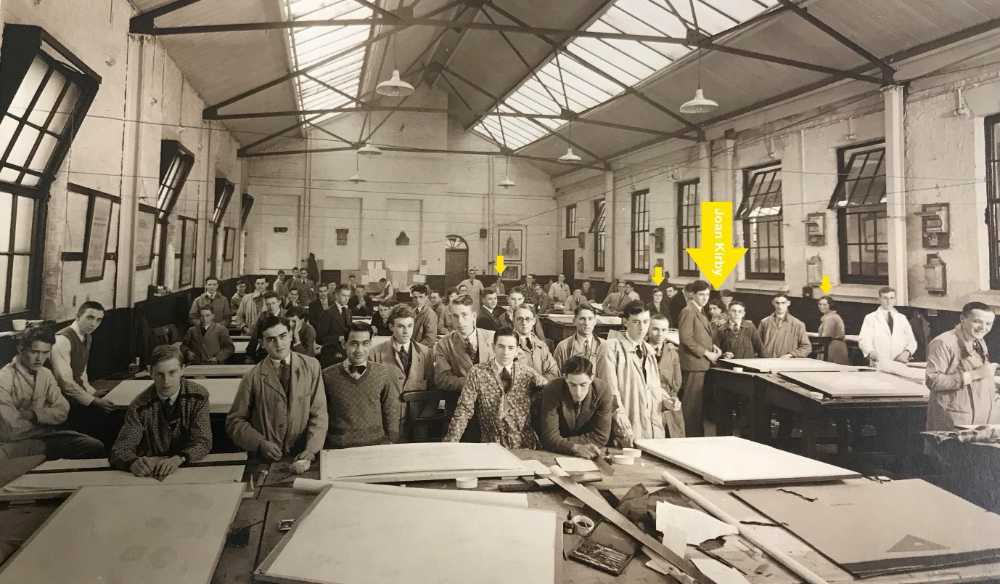 Vintage photograph inside a large teaching studio. Male students inoverals,, shirts and ties stand leaning on drwaing boards. Arrows point out three female students amongst them.