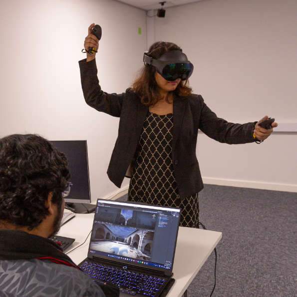 A smartly dressed woman wearing a set of VR goggles and holding handsets in outstretched hands stands navigating a virtual environment. In front of her a man looks at the screen of a laptop showing the VR environment.