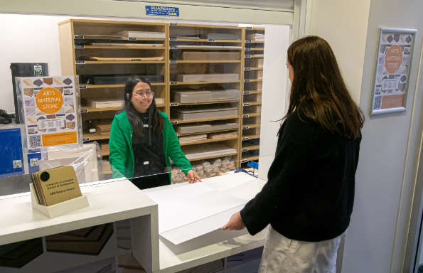 A woman holding a large sheet sheet of card in front of a counter. Behind the counter another woman is smiling.