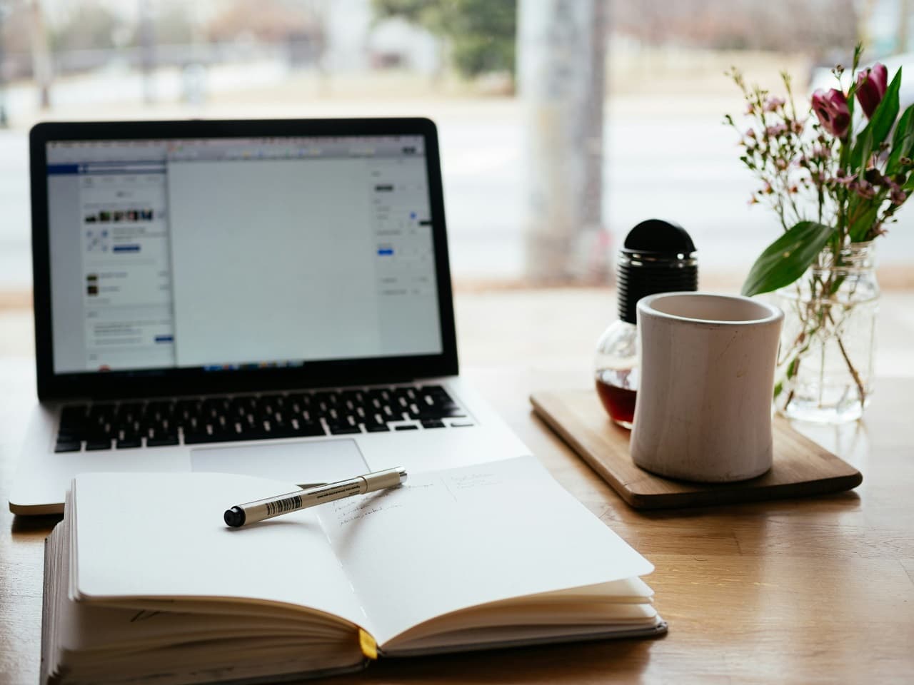 A laptop is open on a table with a mug next to it and a notepad with pen in front of it.