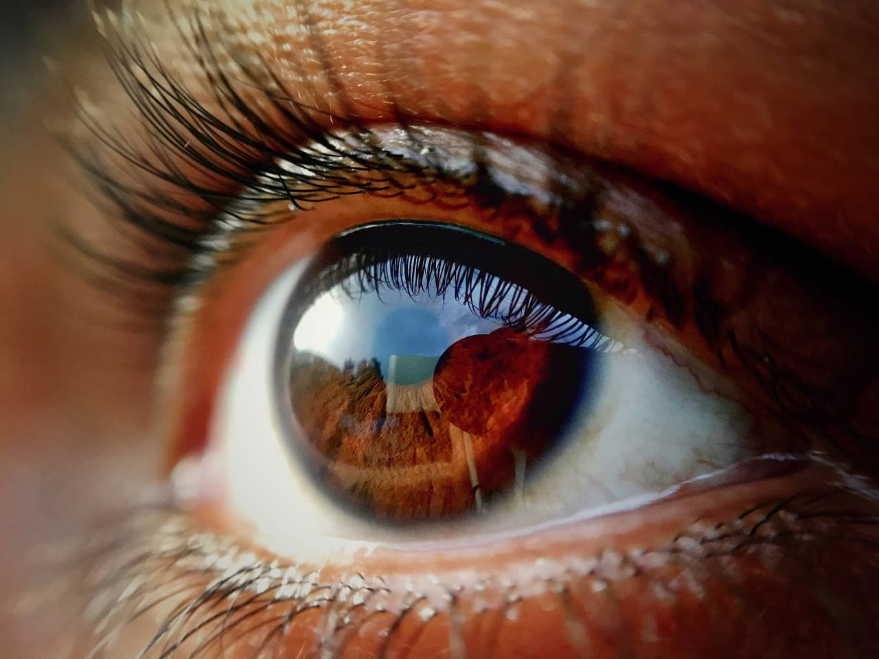 A close up shot of a person's eye looking upwards with the sky and buildings reflected in it.