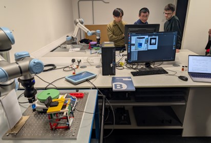 A diverse group of people gathers around a desk, observing a robot placed in the center of the workspace.