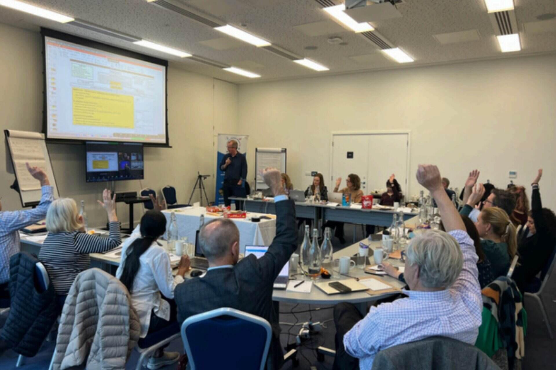 A group of peoeple around a table facing a presentation with their hands up in the air.