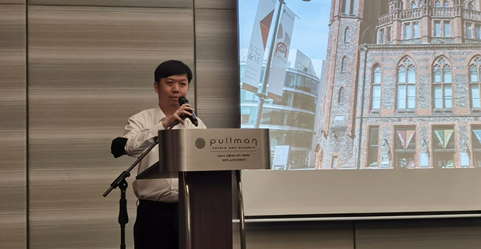 A person stood behind a lectern, presenting in front of a projector screen.