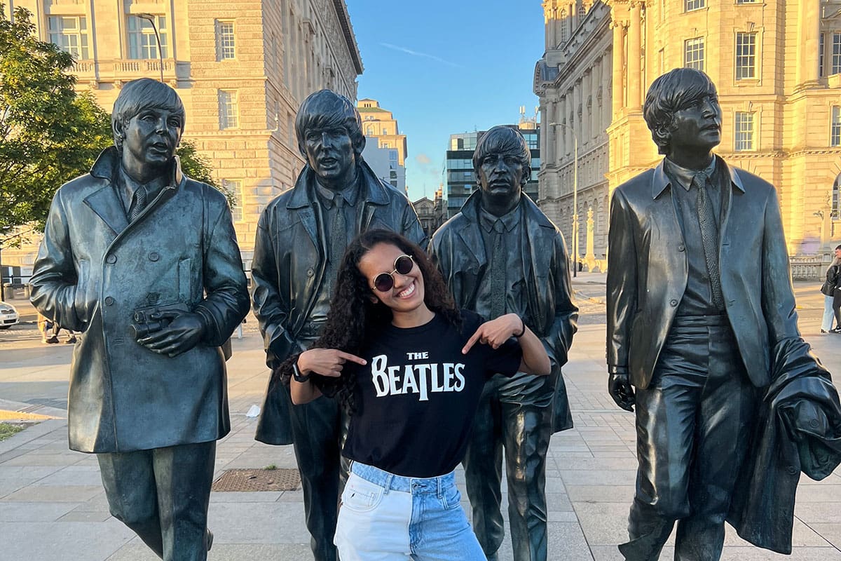 A female student wears a Beatles t-shirt and stands in front of The Beatles statues in Liverpool.