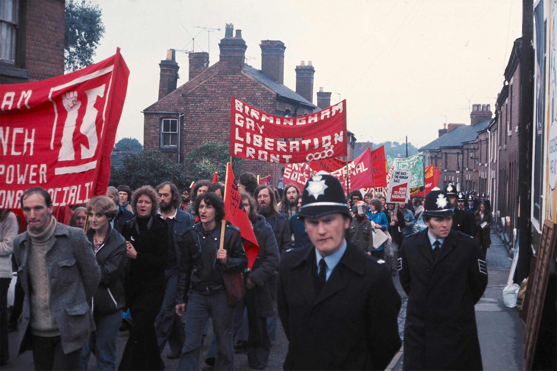 Group of people marching down a street holding signs and protesting for gay liberation, while police officers walk down the side