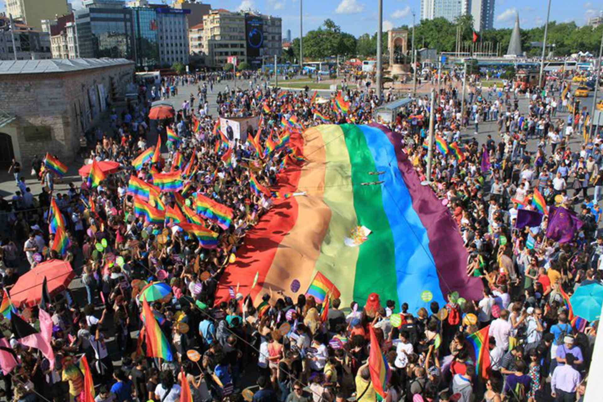 Large crowd of people at gay pride event carrying a huge rainbow flag and others carrying and marching with smaller rainbow flags